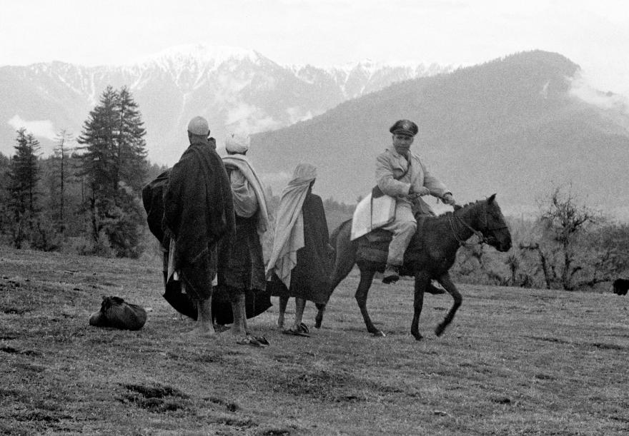 A UN officer on horseback speaks with three locals in cloaks on a grassy field, with snow-capped mountains and tall trees in the background.